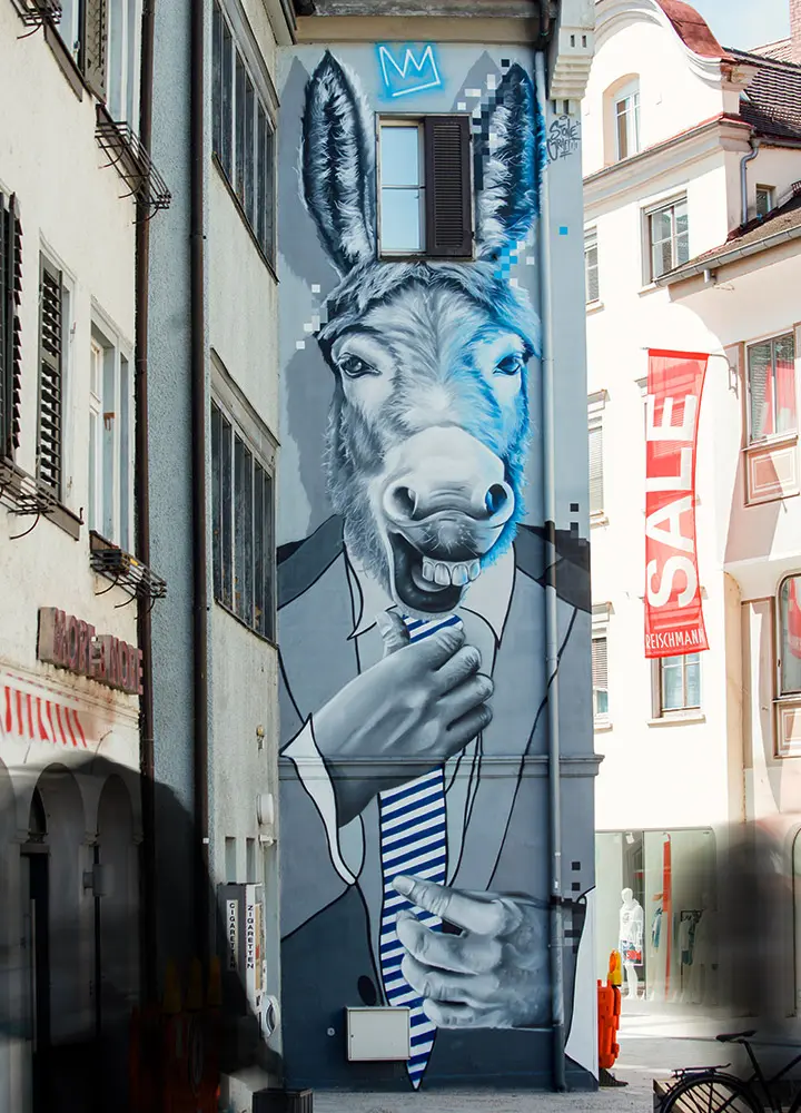 Ein großes Graffiti an der Wand eines schmalen Gebäudes zeigt den Kopf eines Esels, der einen Anzug und eine blau-weiß gestreifte Krawatte trägt. Der Esel hat einen schelmischen Gesichtsausdruck und hält die Krawatte mit einer Hand fest. Über dem Esel ist eine Krone gemalt. Rechts im Bild ist ein rotes Banner mit der Aufschrift 'SALE' an einem benachbarten Gebäude zu sehen.