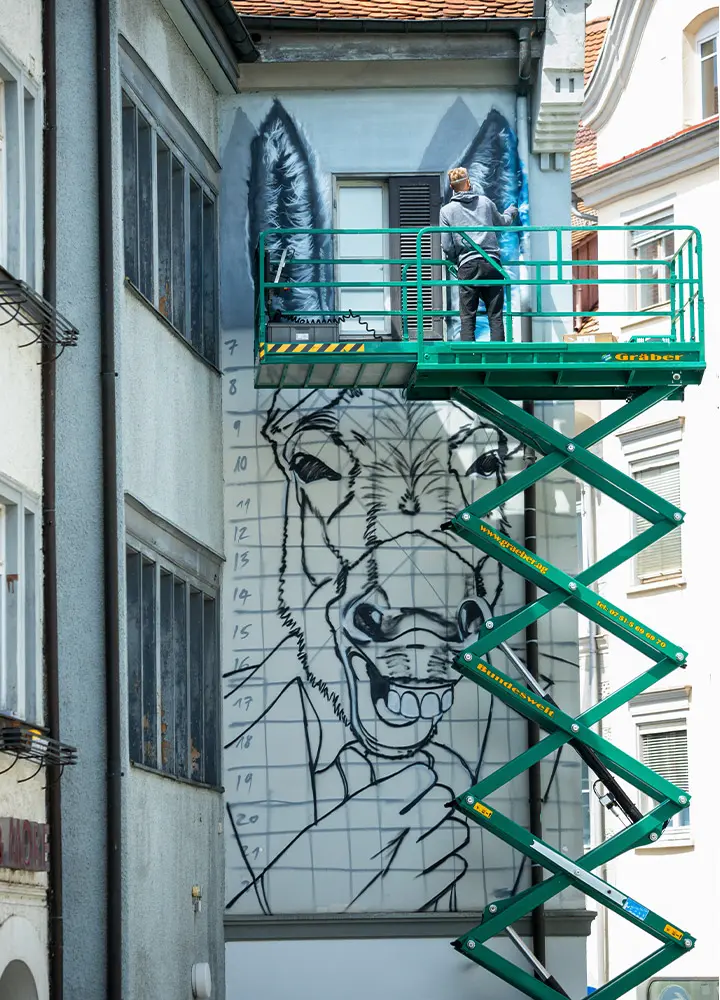 Der Künstler Stone Graffiti arbeitet auf einer Hebebühne an einem großen Graffiti, das einen Esel in einem Anzug zeigt. Der Künstler sprüht Details auf die Ohren des Esels, während das Bild auf eine gerasterte Wand aufgetragen wird. Das Bild zeigt die Fortschritte des Kunstwerks.