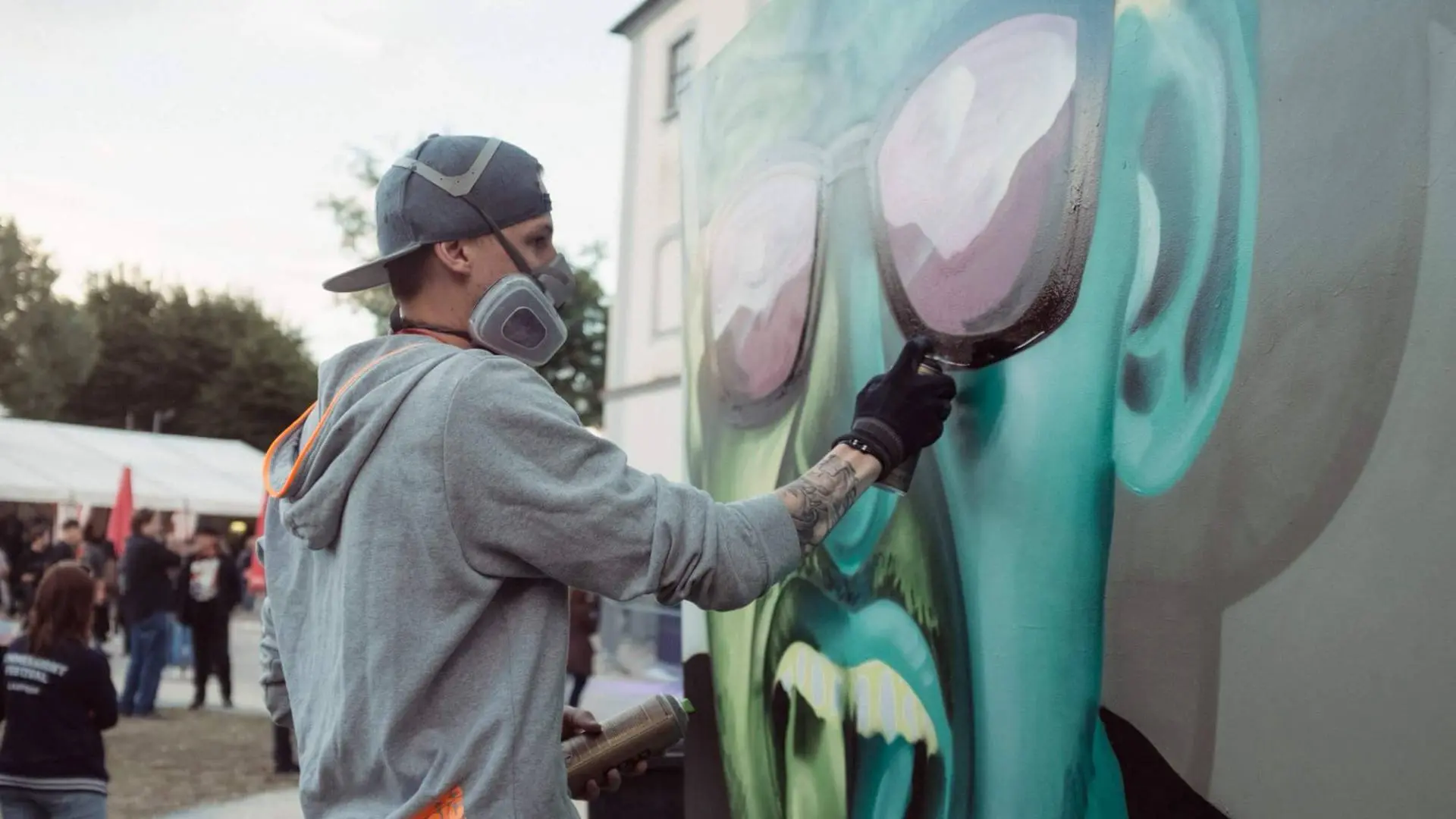 Der Graffiti-Künstler, Philip Walch, ein junger blonder Mann steht vor einer Leinwand unter freiem Himmel. Er sprüht mit einer Graffiti-Spraydose ein Graffiti eines grünen Gesichts mit schwarzer Sonnenbrille an die Leinwand. Dabei trägt er einen grauen Hoodie und einer grau Cap die nachhinter gedreht ist. Seine Nase und sein Mund werden von einer Feinstaubmaske verdeckt. Im Hintergrund sind viele Menschen und ein Festzelt zu erkennen.