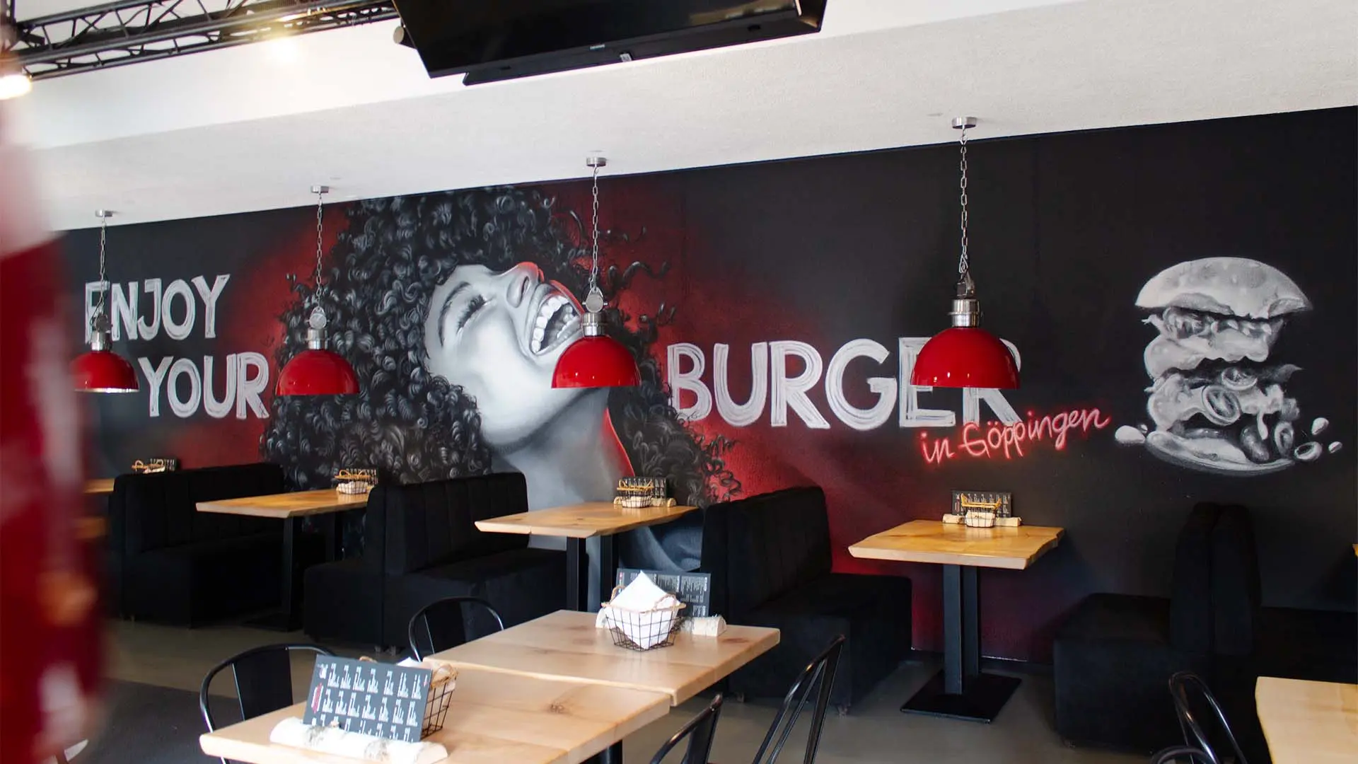 Das Graffiti im Restaurant zeigt das lachende Gesicht einer Frau, umgeben von den Worten 'ENJOY YOUR BURGER' in großen, weißen Buchstaben. Die Wand ist in Schwarz- und Rottönen gestaltet, und über den Tischen hängen rote Lampen. Ein weiteres Graffiti eines Burgers befindet sich rechts neben dem Gesicht, mit dem Zusatz 'in Göppingen' in roter Schrift.